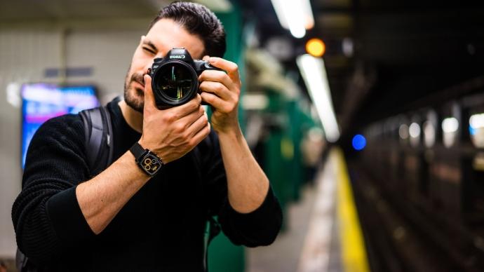 a portrait of Andreas Verrios holding a camera up to his eye in the NYC subway